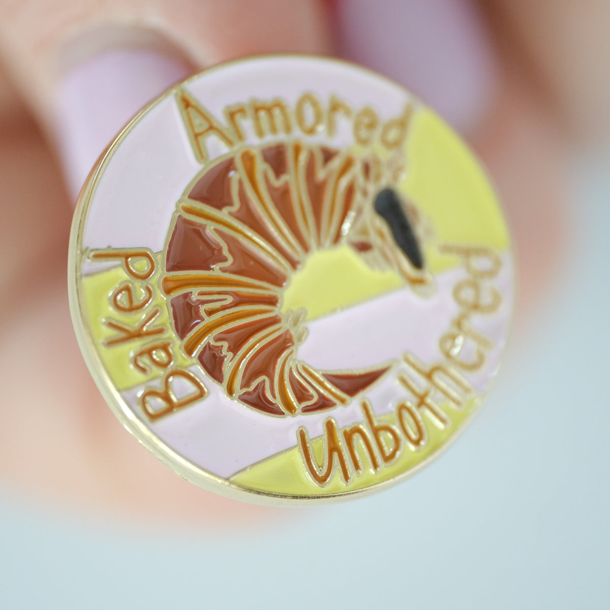 Close-up of croissantadillo enamel pin with an armadillo croissant and Baked, Armored, Unbothered lettering.