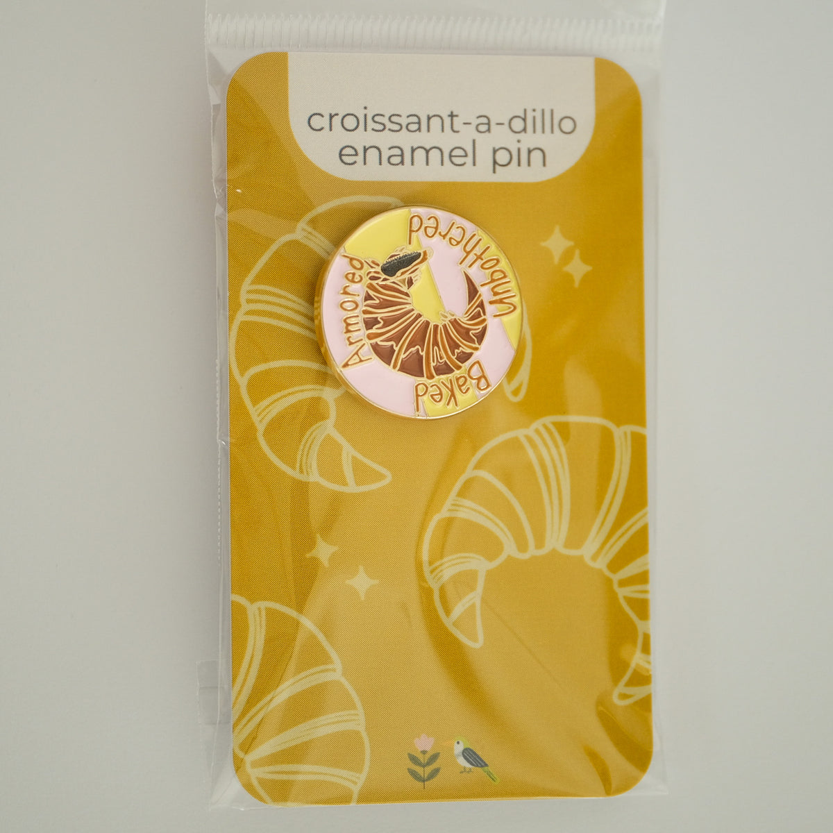 Croissantadillo enamel pin with an armadillo croissant and Baked, Armored, Unbothered lettering on its product backing card.