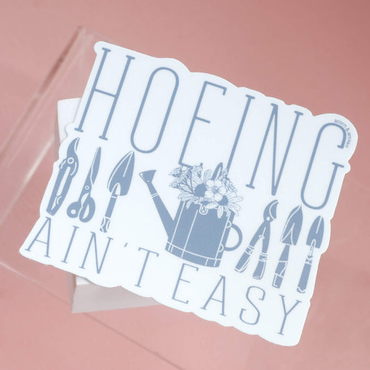 Light blue background of sticker with fun garden themed design. In darker blue the sticker says hoeing ain't easy with vintage garden tools, watering can, and flowers.