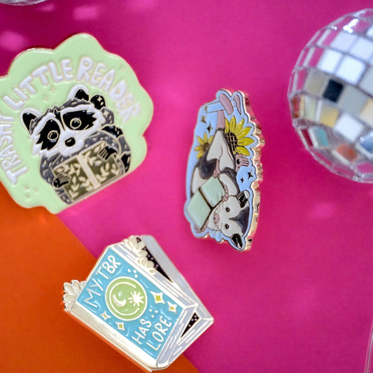 Colorful Acorn & Artisan enamel pins for book lovers, including a reading raccoon, reading opossum, and My TBR Has Lore design