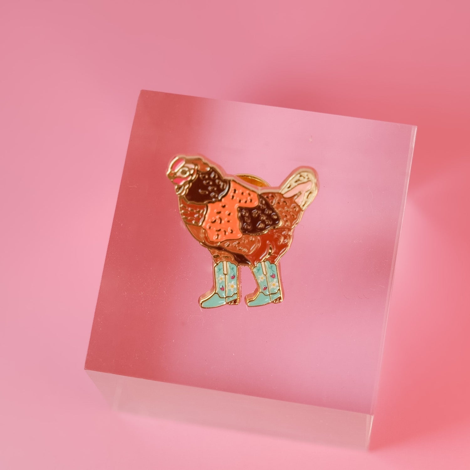 A colorful enamel keychain featuring a stylized chicken in orange and brown with light blue rain boots, attached to a gold-tone clasp and a small black circular charm. The keychain is styled on a pink and orange background with disco ball props.