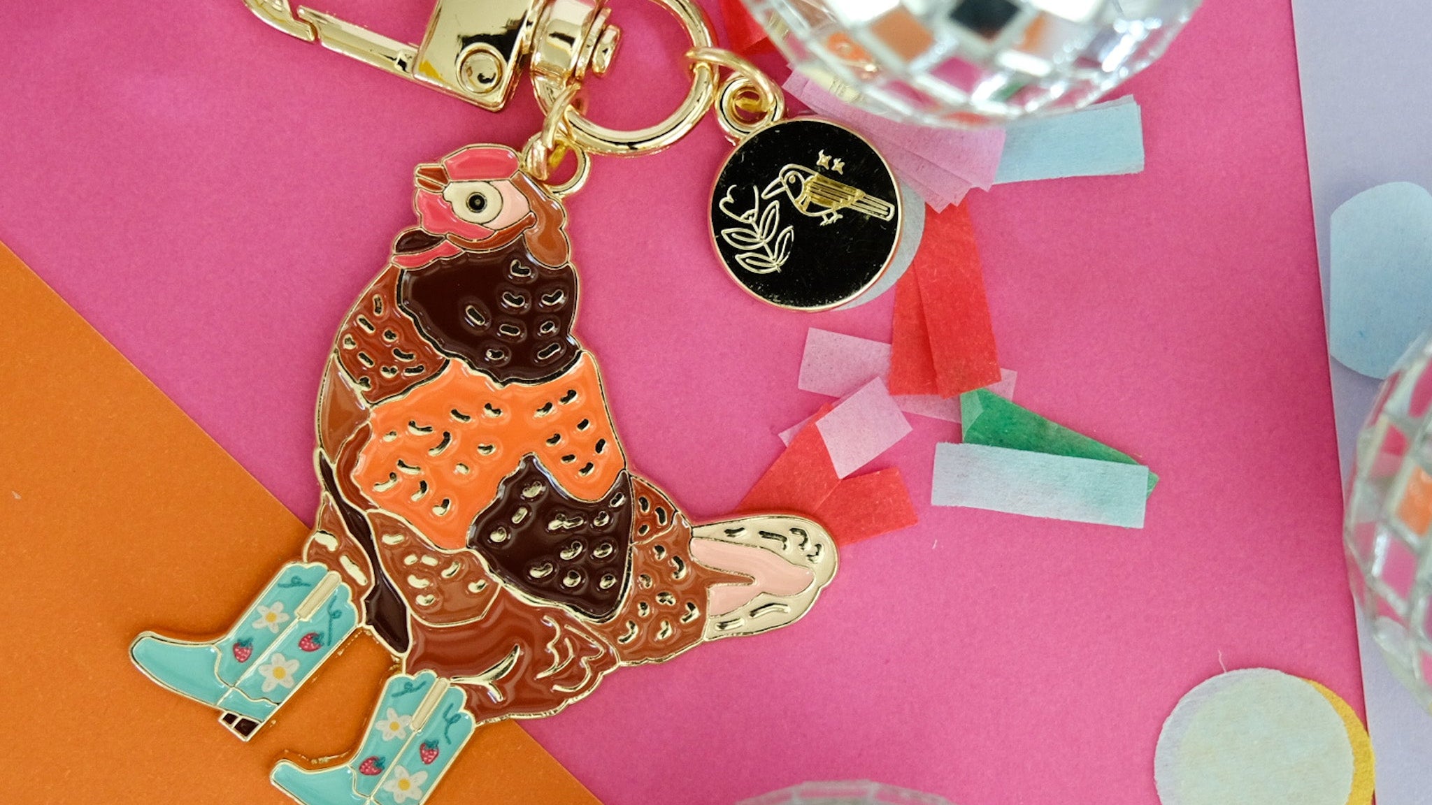 A colorful enamel keychain featuring a stylized chicken in orange and brown with light blue rain boots, attached to a gold-tone clasp and a small black circular charm. The keychain is styled on a pink and orange background with disco ball props.