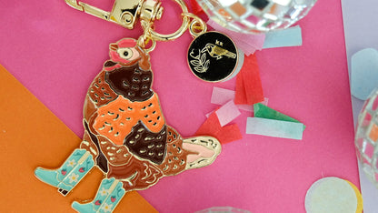 A colorful enamel keychain featuring a stylized chicken in orange and brown with light blue rain boots, attached to a gold-tone clasp and a small black circular charm. The keychain is styled on a pink and orange background with disco ball props.