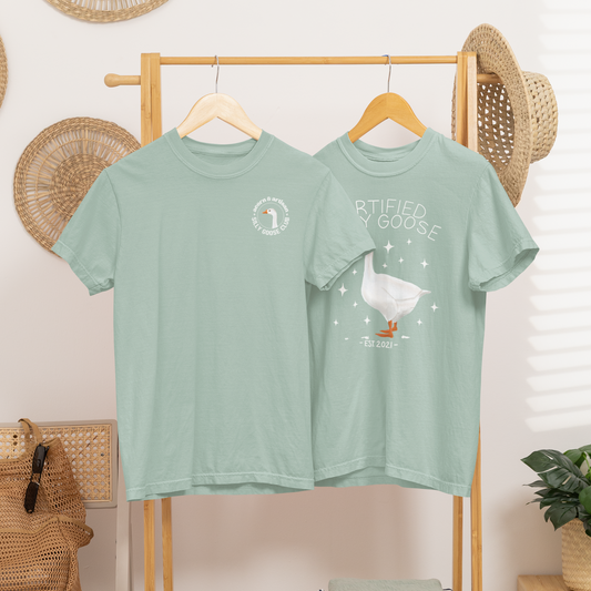 Two green t-shirts on a wooden rack with boots and a plant in the background.